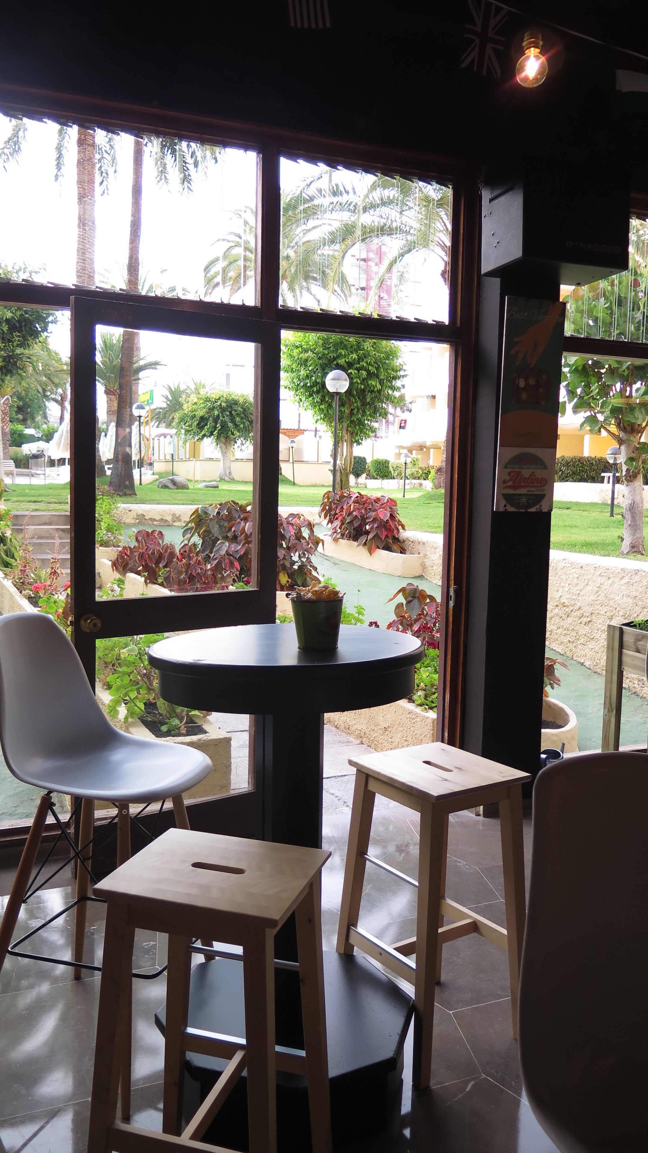Interior de restaurante con una mesa y dos sillas, ventanas amplias con vista a un jardín y palmeras.