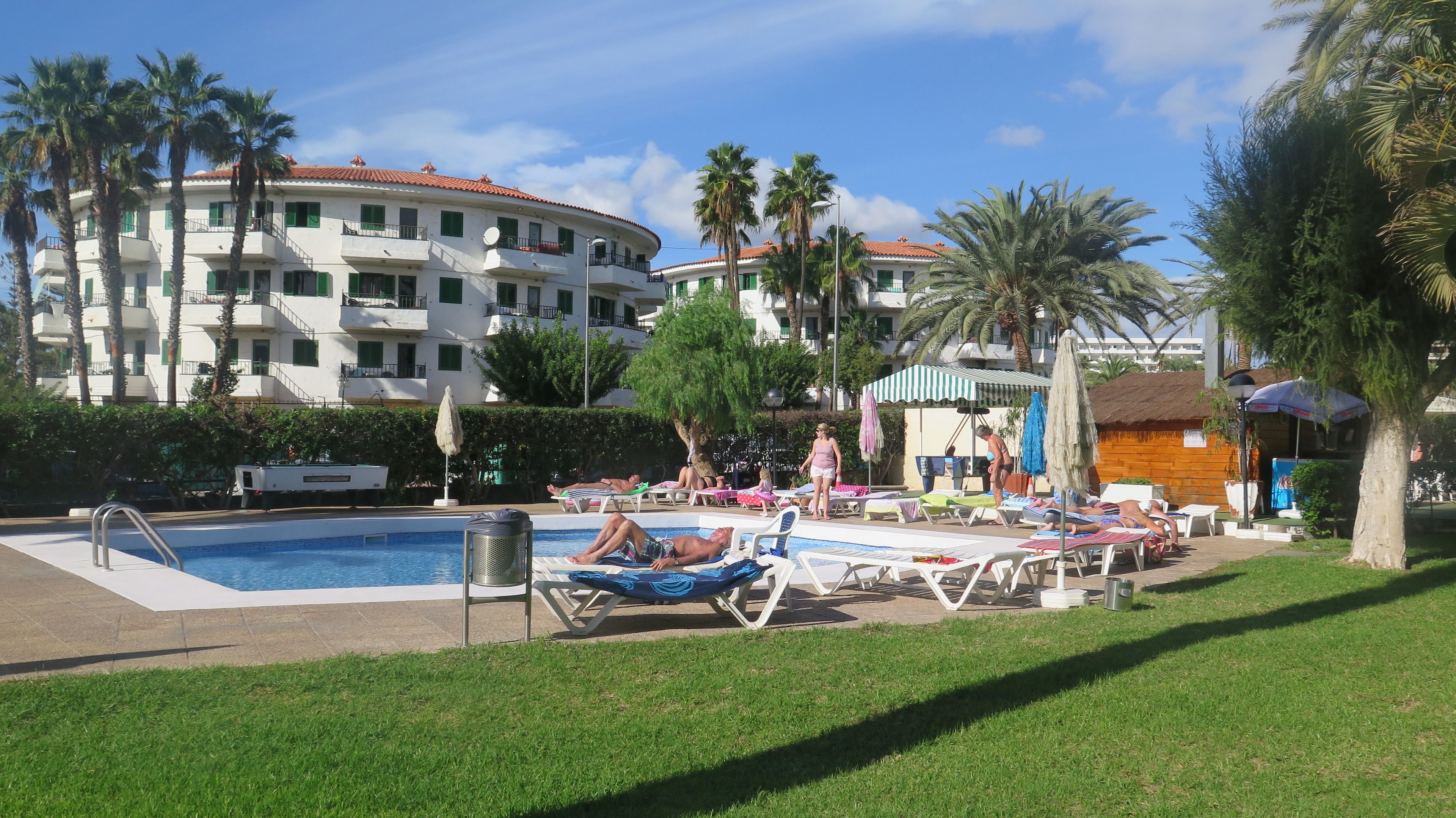 A pool area with lounge chairs, a swimming pool, and a building with balconies and palm trees in the background.