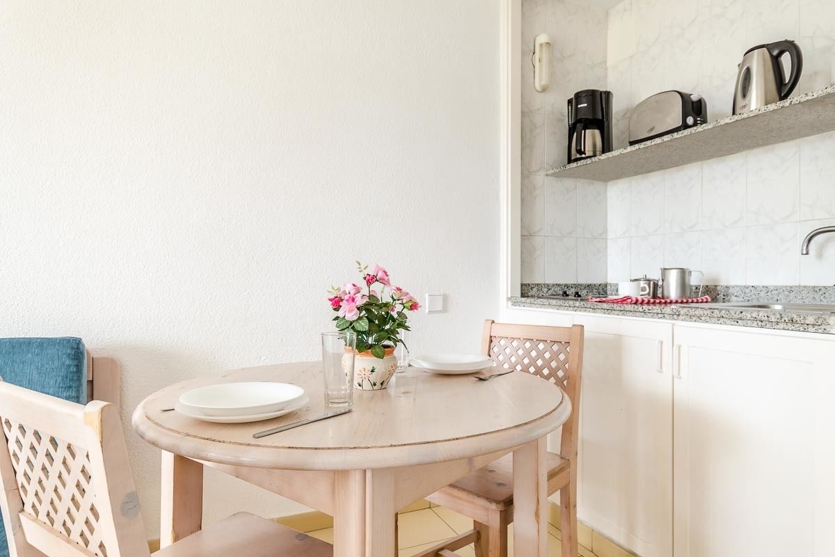 Una pequeña cocina con una mesa redonda de madera, dos sillas, un florero con flores y una cafetera en la alacena.