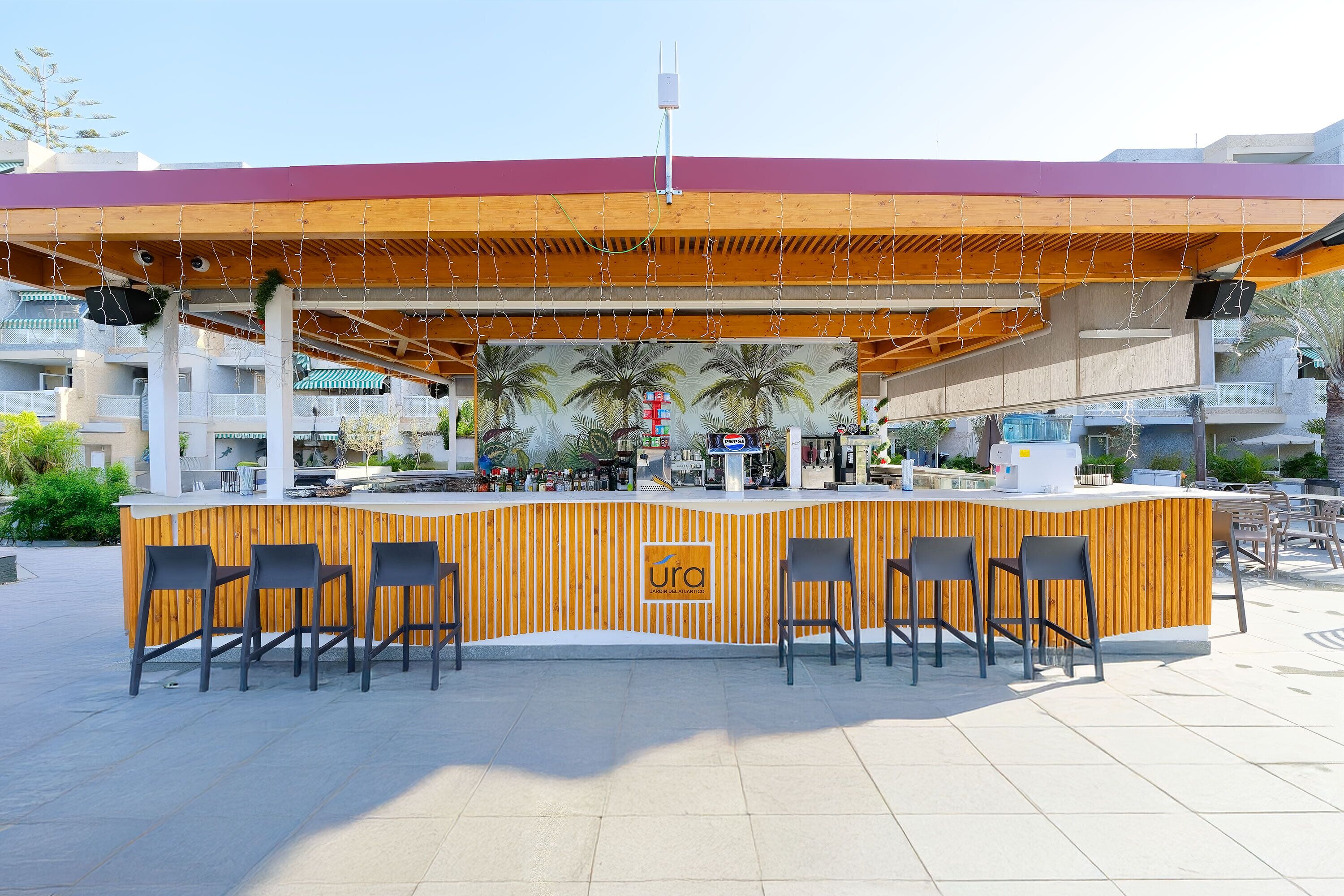 Un área de bar al aire libre con mostrador de madera, taburetes de bar y un mural de palmeras.