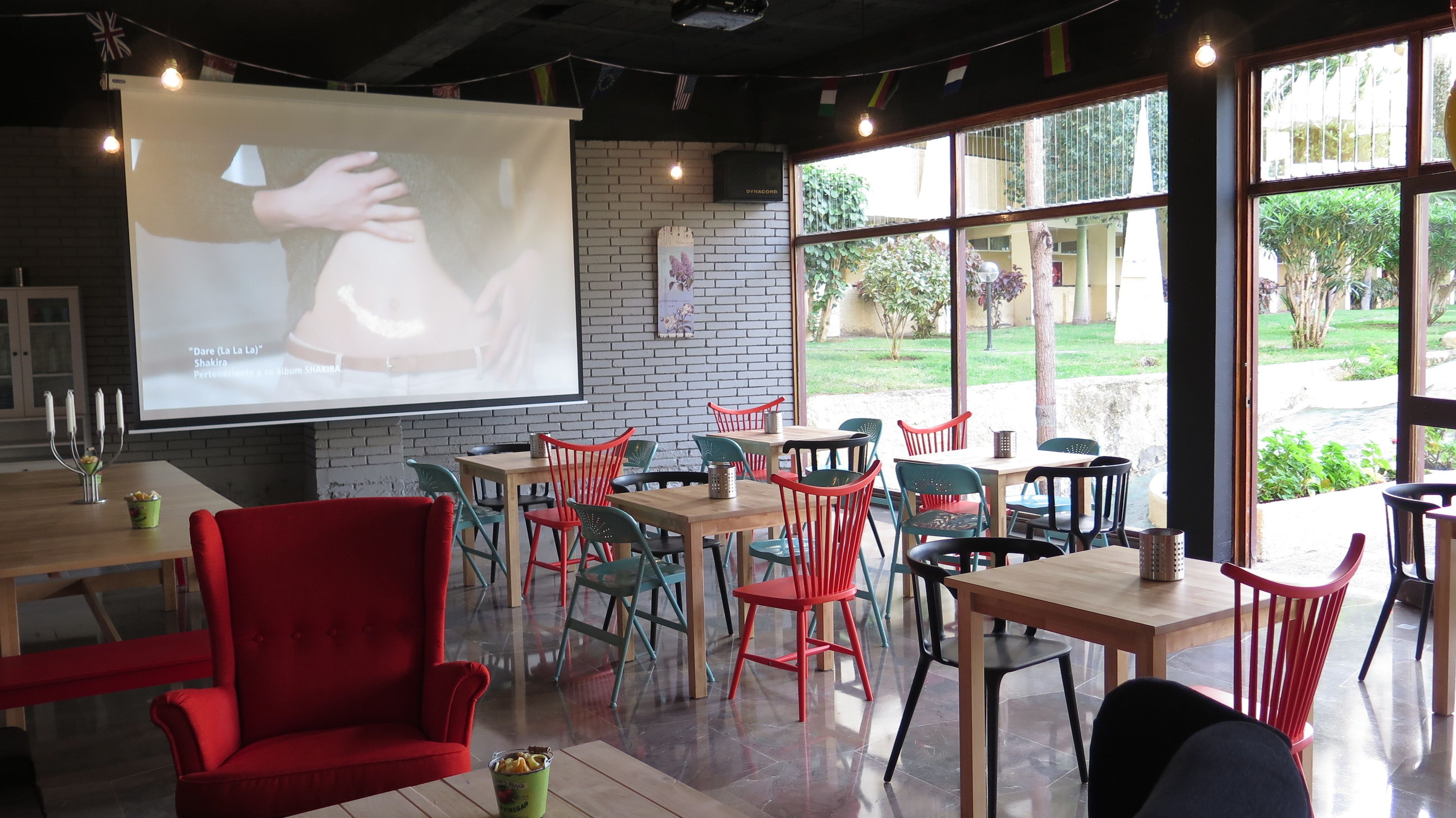 A cozy restaurant with a projector screen displaying a dance performance, wooden tables, and red chairs.