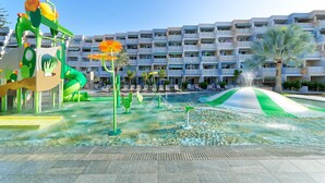A water park with slides and play structures, a pool with a wave feature, and a backdrop of a multi-story building with balconies.