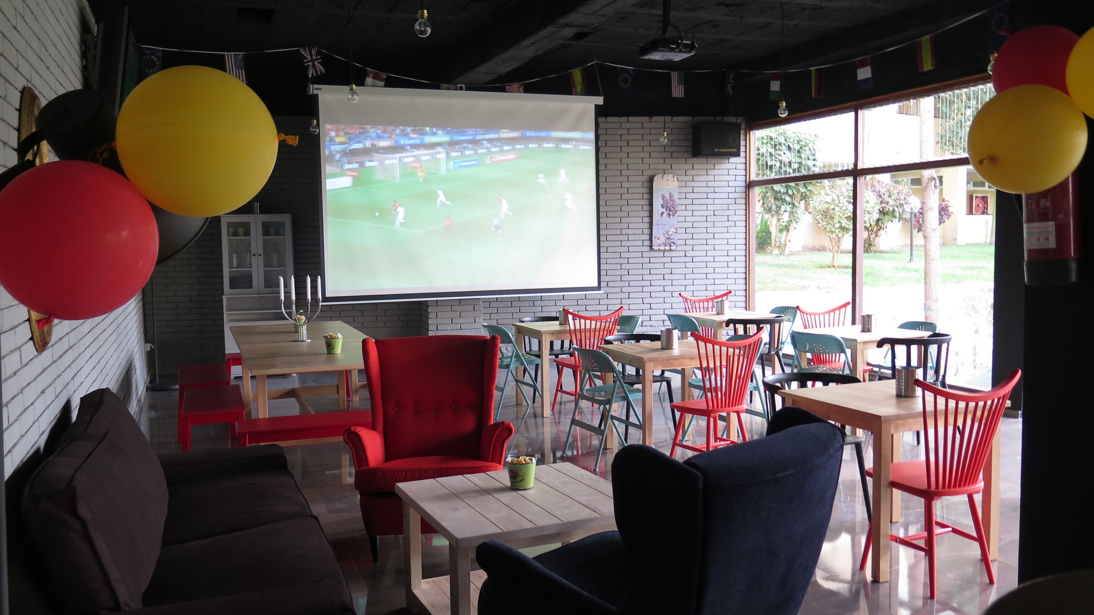 Una habitación acogedora con una pantalla grande mostrando un partido de fútbol, globos rojos y amarillos, y una pared de ladrillo.