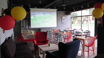 Una habitación acogedora con una pantalla grande mostrando un partido de fútbol, globos rojos y amarillos, y una pared de ladrillo.