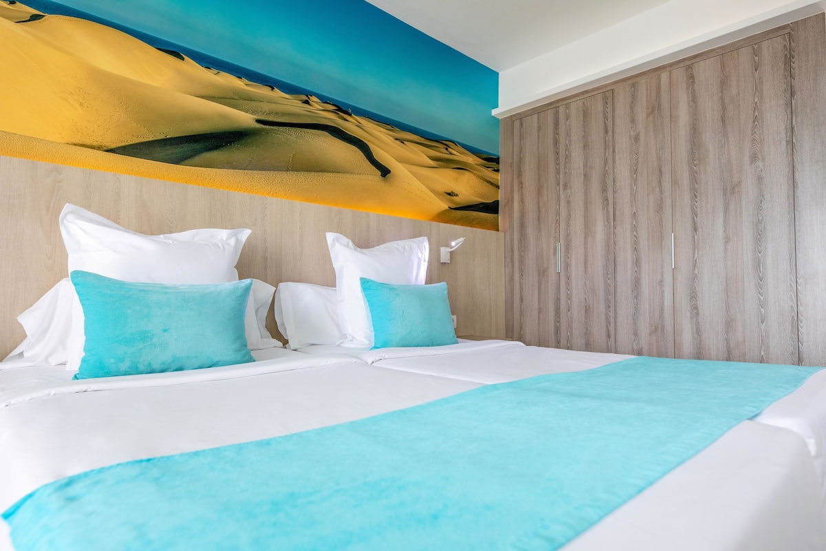 Ein Hotelzimmer mit Bett, Kissen, Decke, einem Holzschrank und einem Wüstenwandbild.