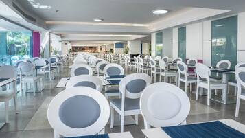 A spacious dining area with white chairs and tables, a visible kitchen area in the background, and large windows allowing natural light.