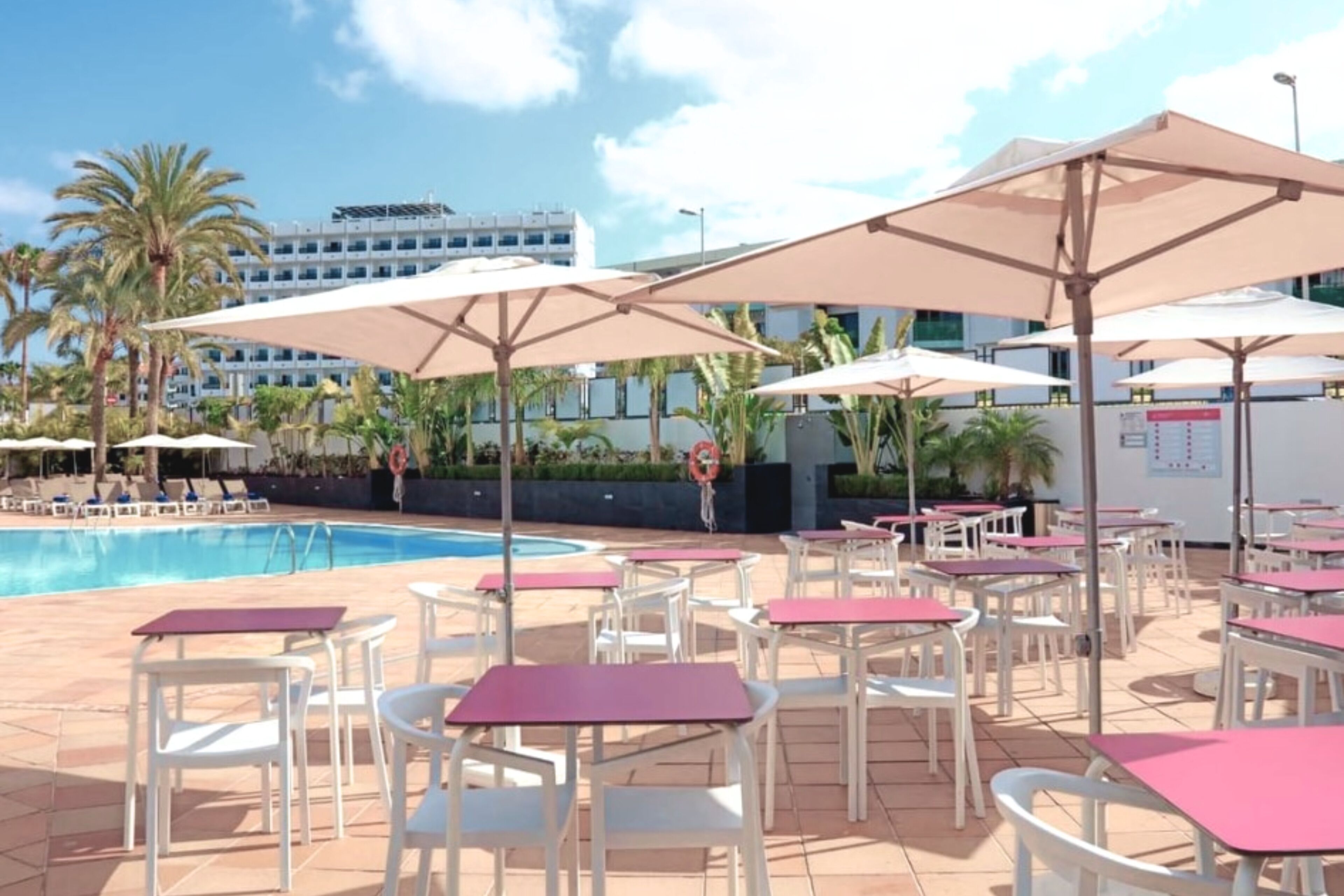 Un espace aménagé au bord de la piscine, avec des tables et des chaises blanches, des parasols beige et une piscine à l’eau d’un bleu limpide.