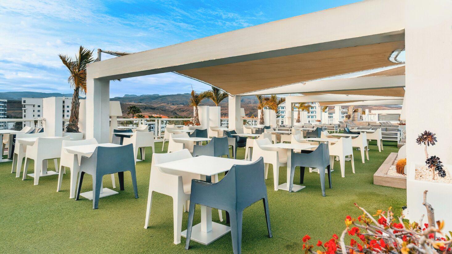 An outdoor dining area with white tables and chairs, covered by a canopy, and a view of buildings and hills in the background.