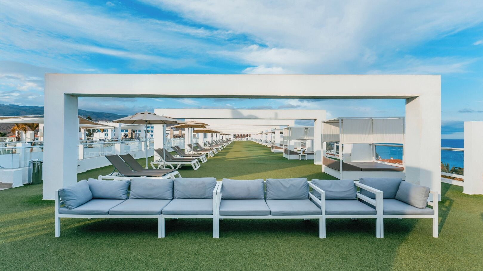 Zona de lounge en la azotea con una fila de tumbonas y un sofá blanco, con vistas a un puerto deportivo y al mar.