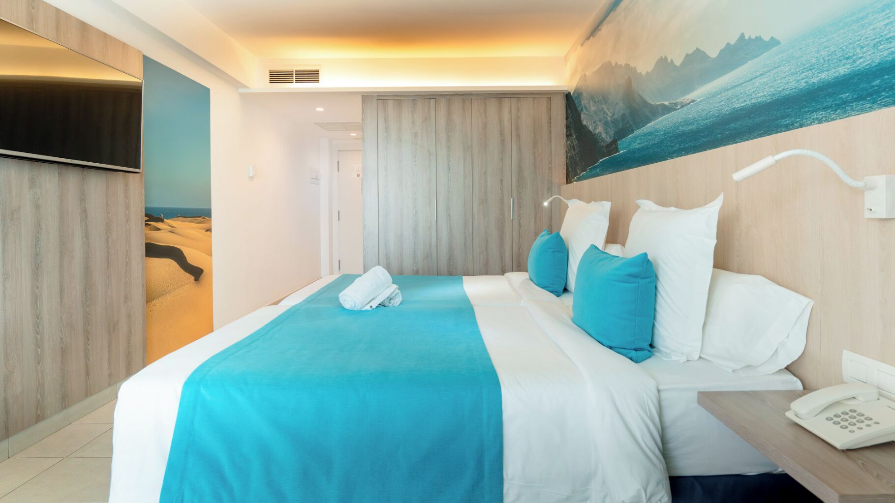 A hotel room with a bed, a nightstand with a phone, and a wall-mounted TV.