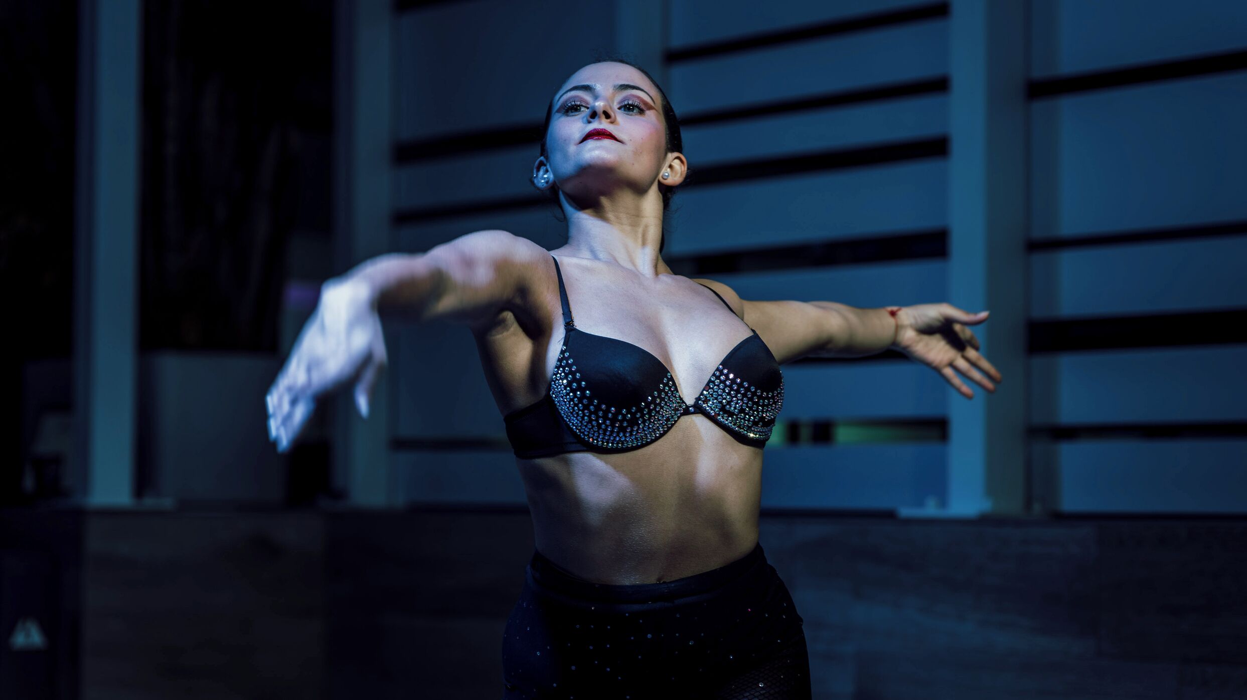 A person in a black outfit with a studded bra, performing a dance in front of a building with horizontal lines.