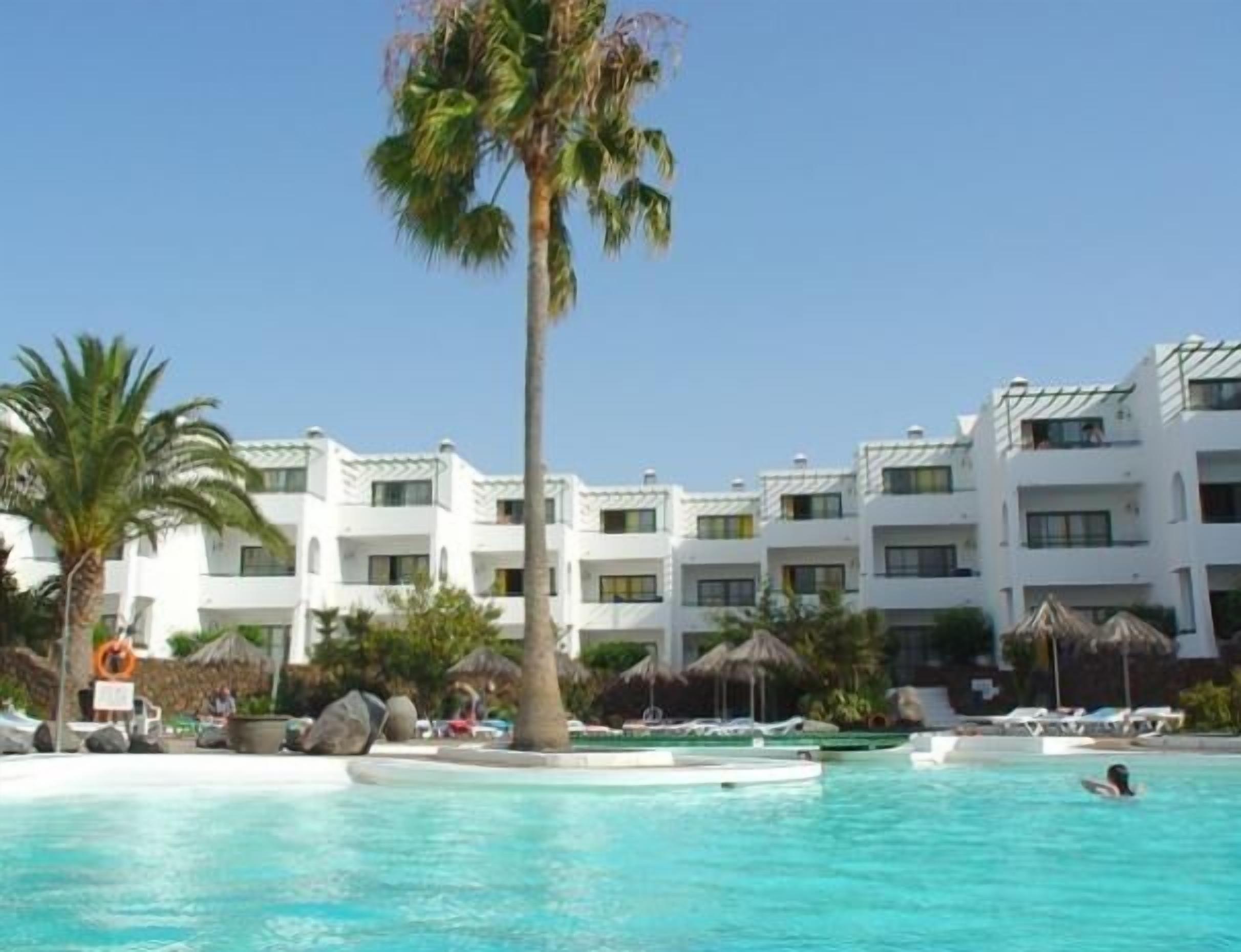 Un complexe hôtelier doté d’une piscine, de palmiers et d’un bâtiment blanc orné de nombreux balcons.