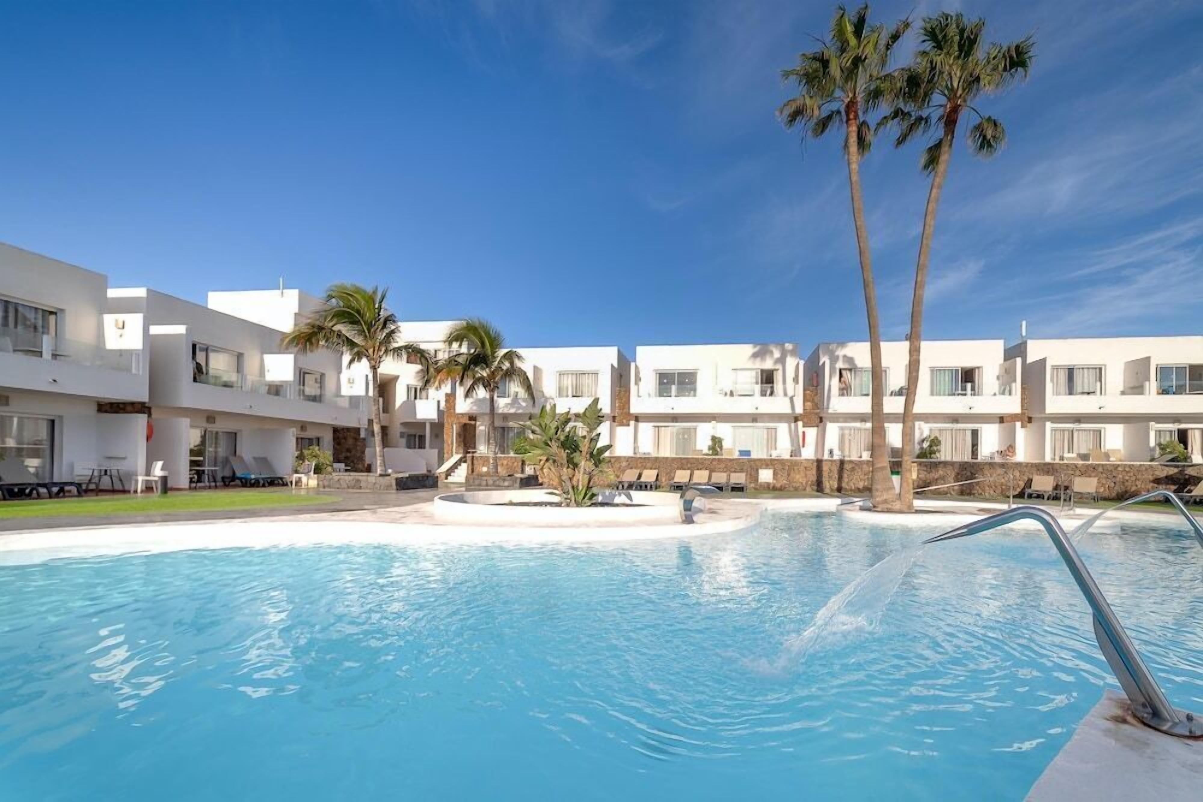 A swimming pool with clear blue water, surrounded by modern buildings with balconies and palm trees.