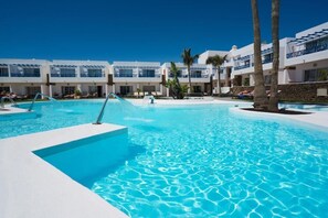 Un espace piscine d’un complexe hôtelier avec une eau d’un bleu limpide, des palmiers et des bâtiments blancs modernes dotés de balcons.