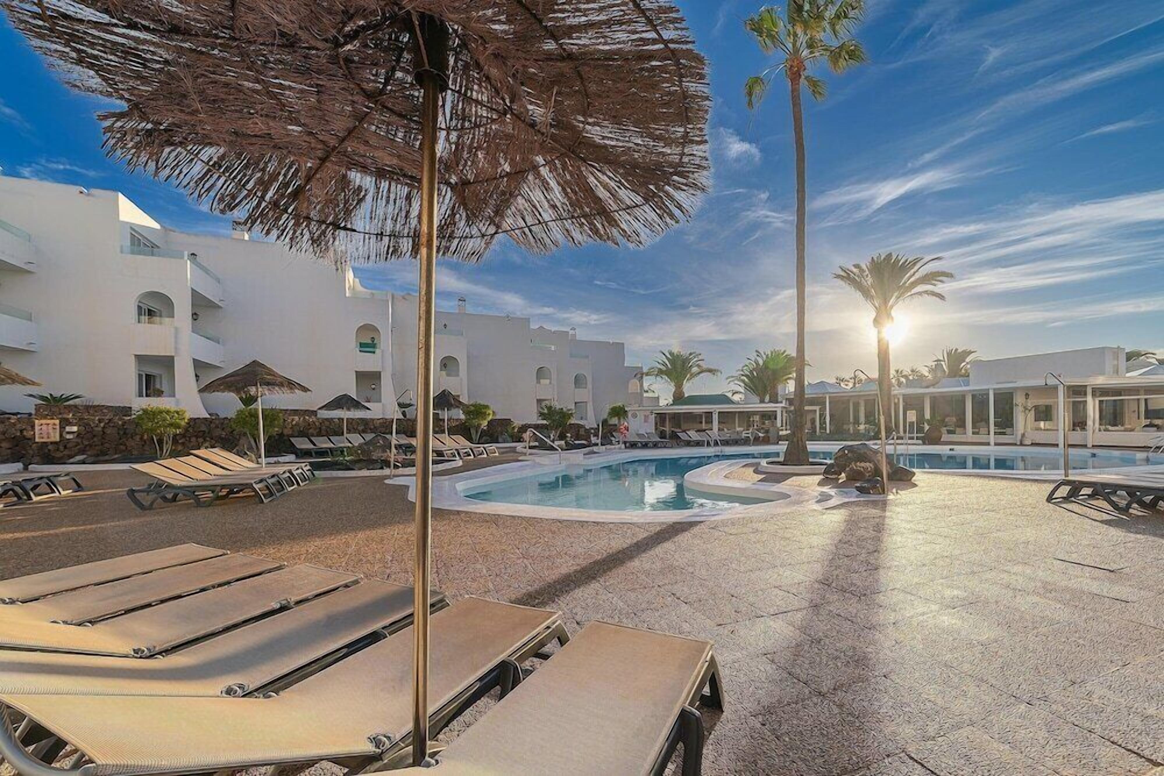 Área de piscina en un resort, con tumbonas, sombrillas y palmeras.