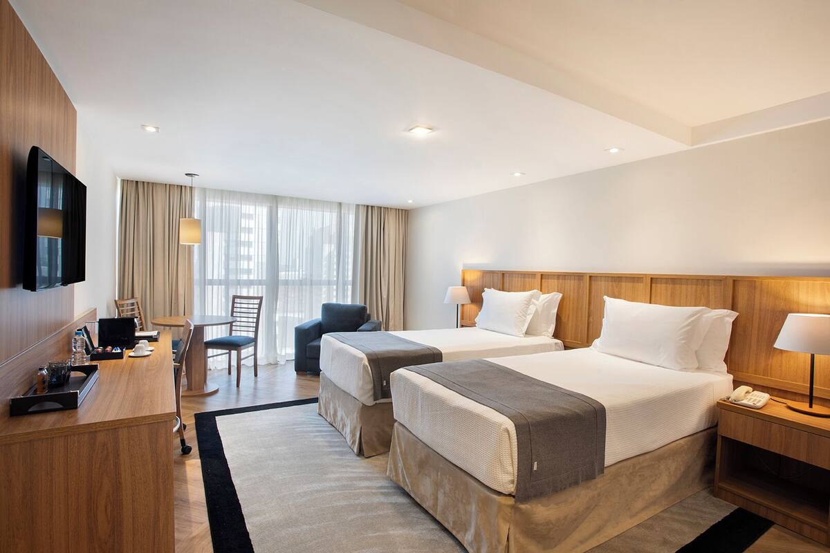 A hotel room with two beds, a desk, a chair, a TV, and a window with curtains.