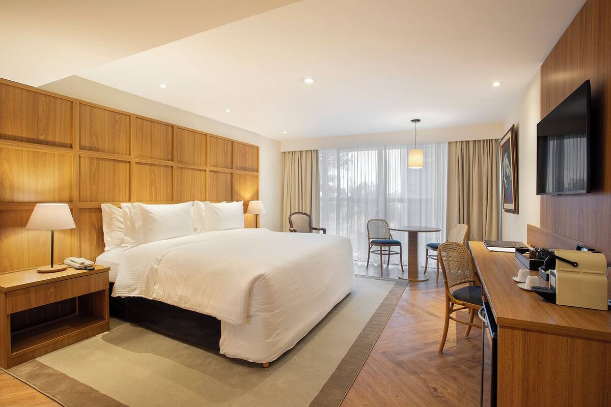 A hotel room with a large bed, bedside tables, a desk with chairs, a TV, and a window with curtains.