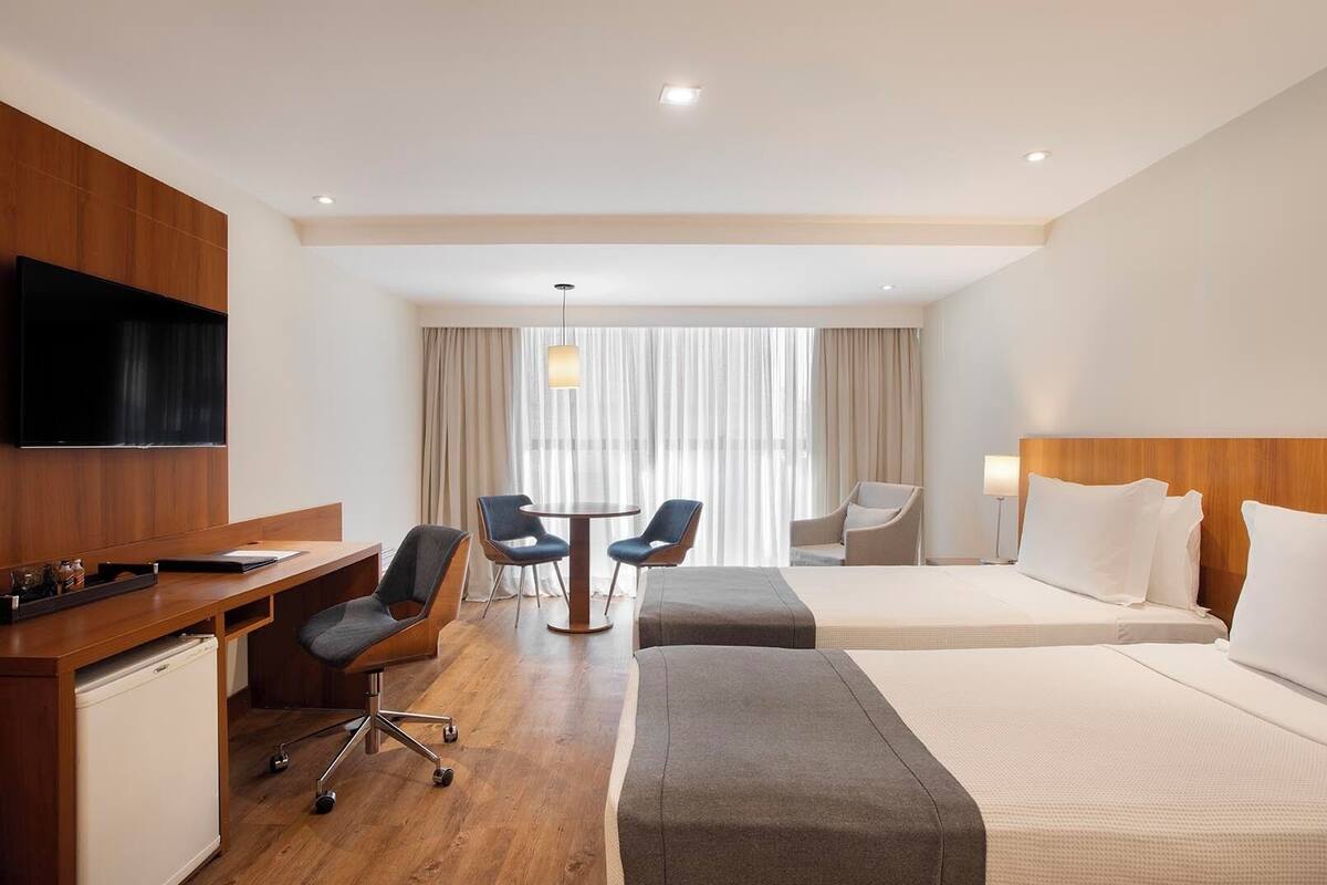 A hotel room with a bed, a desk, a TV, a chair, a table, and a window with curtains.