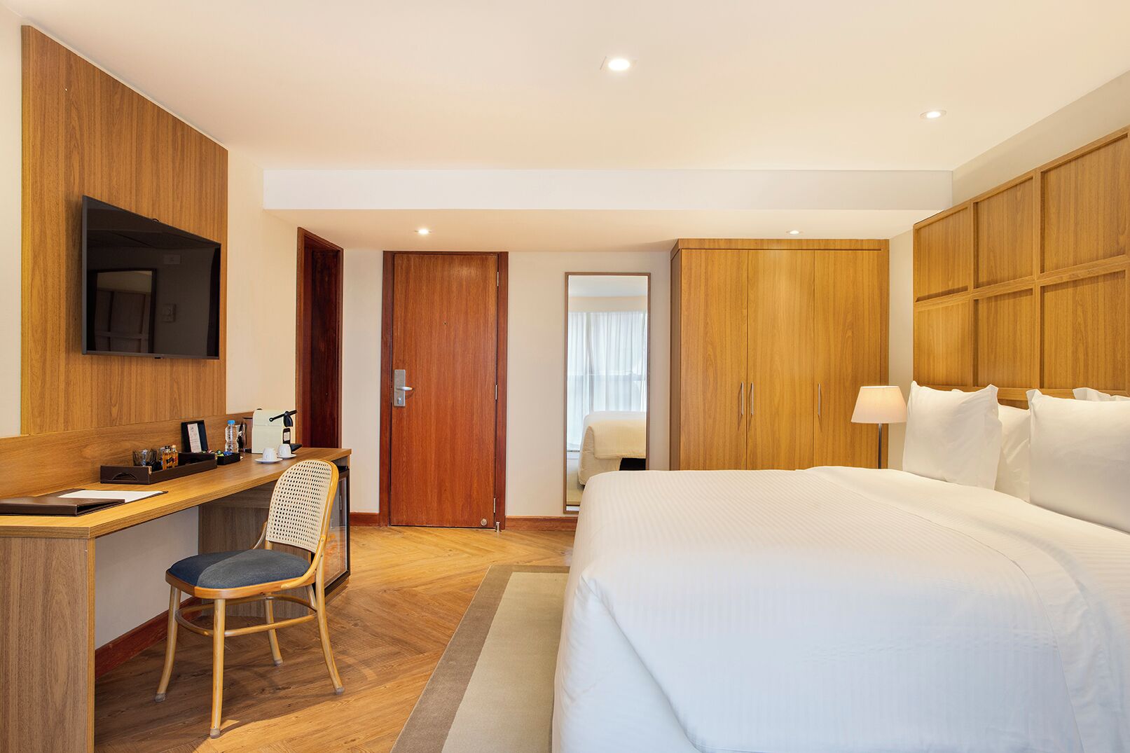 Quarto de hotel com cama, escrivaninha, cadeira, guarda-roupa e televisão na parede.