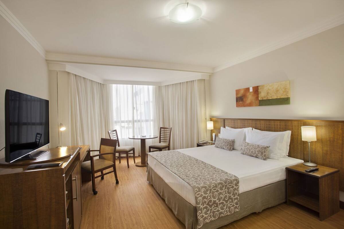 A hotel room with a bed, a desk with a TV, two chairs, a small table, and a nightstand with a lamp.
