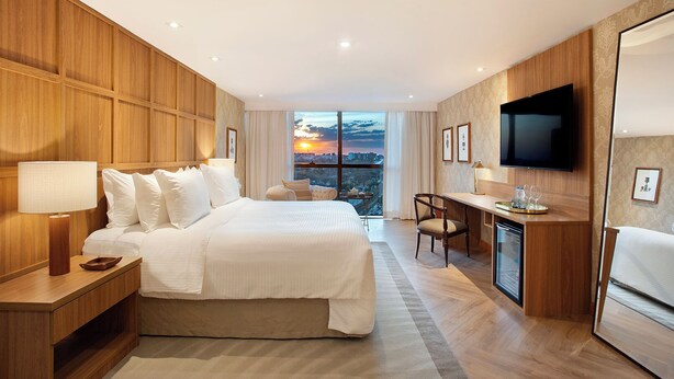 Habitación de hotel con una cama grande, un escritorio, un televisor y vistas a la ciudad al atardecer.