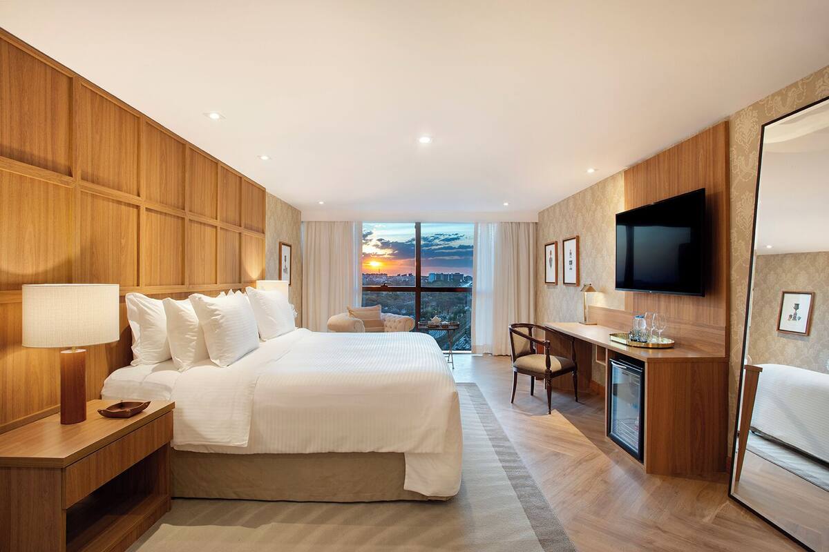 A hotel room with a large bed, a desk, a TV, and a view of the cityscape at sunset.
