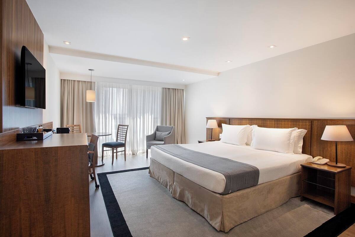 A hotel room with a large bed, a desk, a chair, a TV, and a lamp.