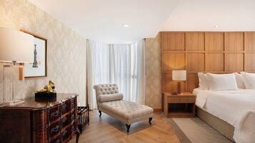 Habitación de hotel con un armario de madera, un sillón capitonado, una mesita de noche con lámpara, una cama grande con ropa de cama blanca y una ventana con cortinas de gasa.