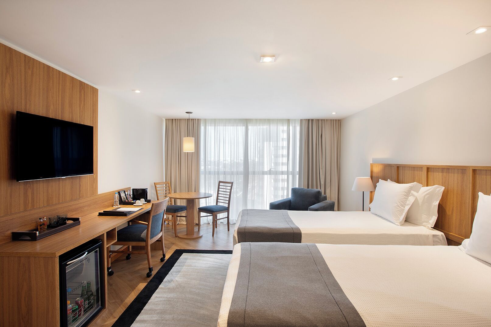 A hotel room with a TV, desk, chairs, a bed, and a window with curtains.