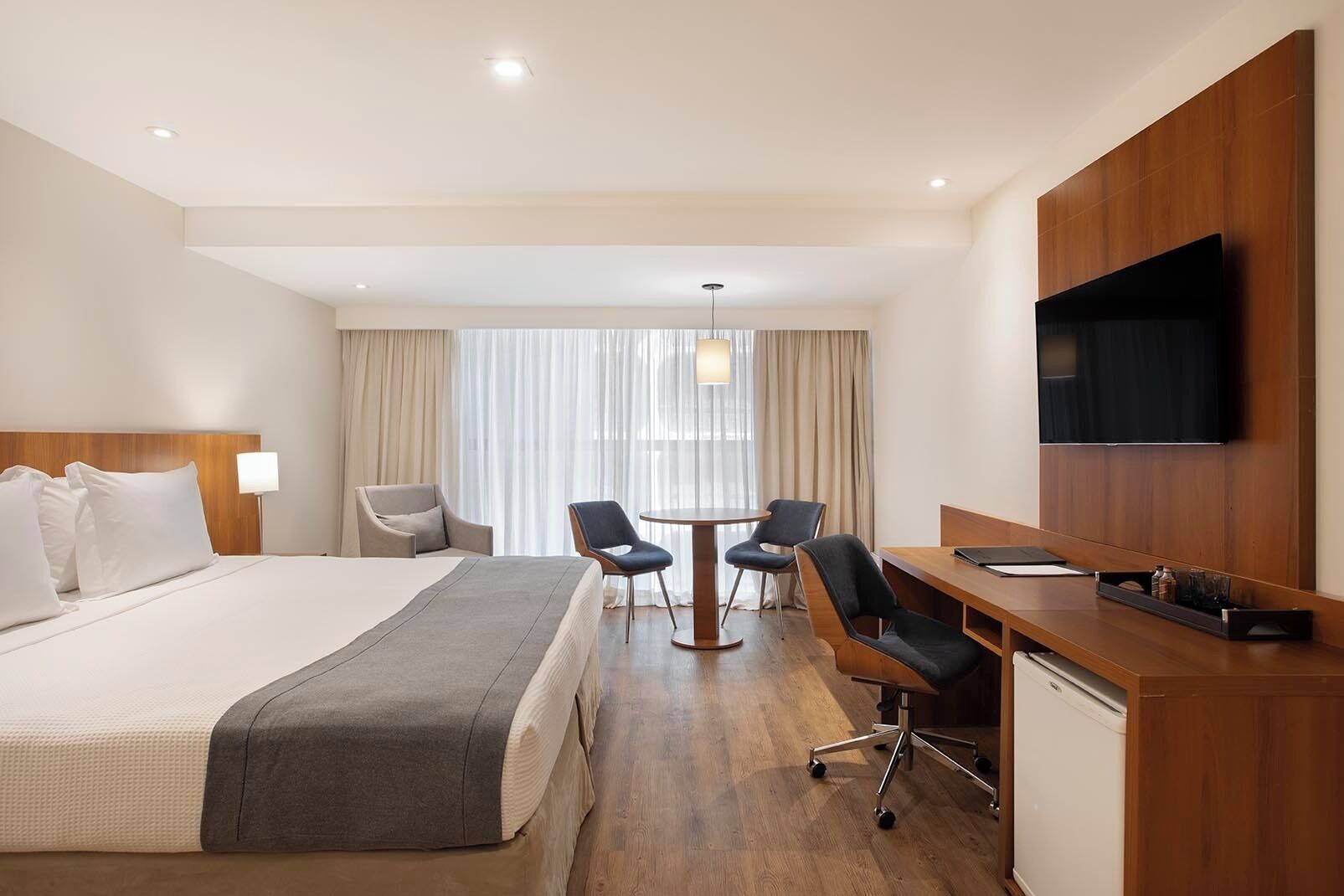 A hotel room with a bed, a desk with a chair, a TV, and a window with curtains.