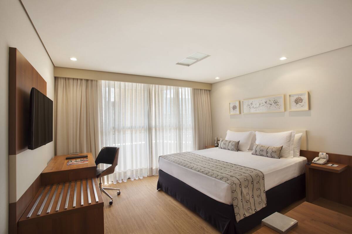A hotel room with a bed, a desk, a chair, a TV, and artwork on the wall.