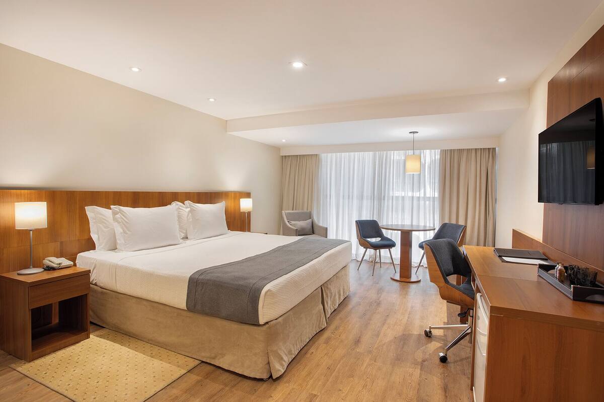 A hotel room with a large bed, bedside tables, a desk with chairs, a TV, and a window with curtains.