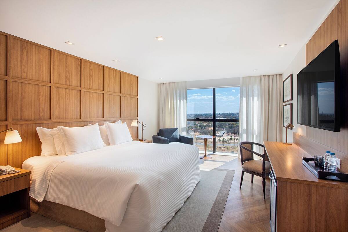 A hotel room with a large bed, a desk with a chair, a TV, and a view of the city through large windows.