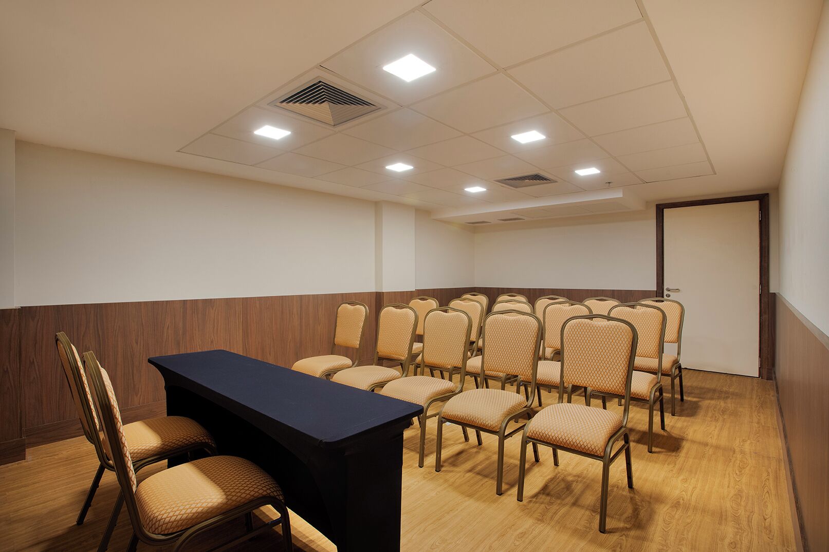 A conference room with a long table and multiple chairs, equipped with ceiling lights and a vent.