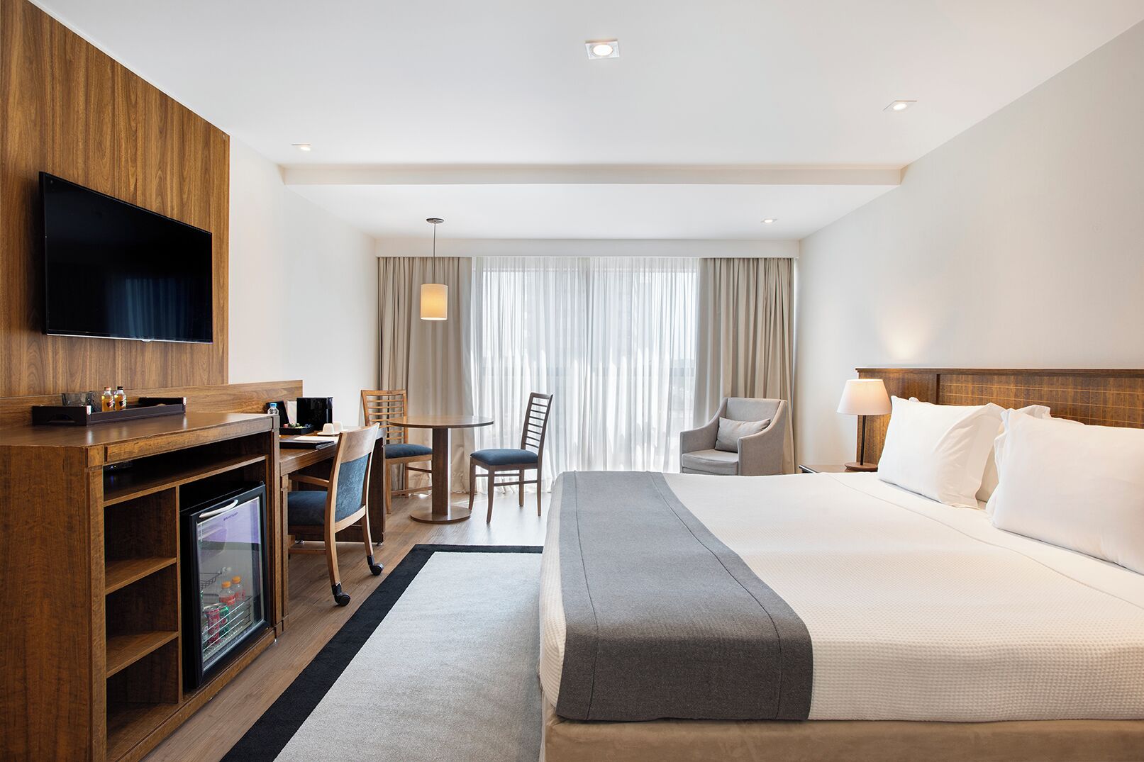 A hotel room with a large bed, a desk with a computer, a TV, and a minibar.