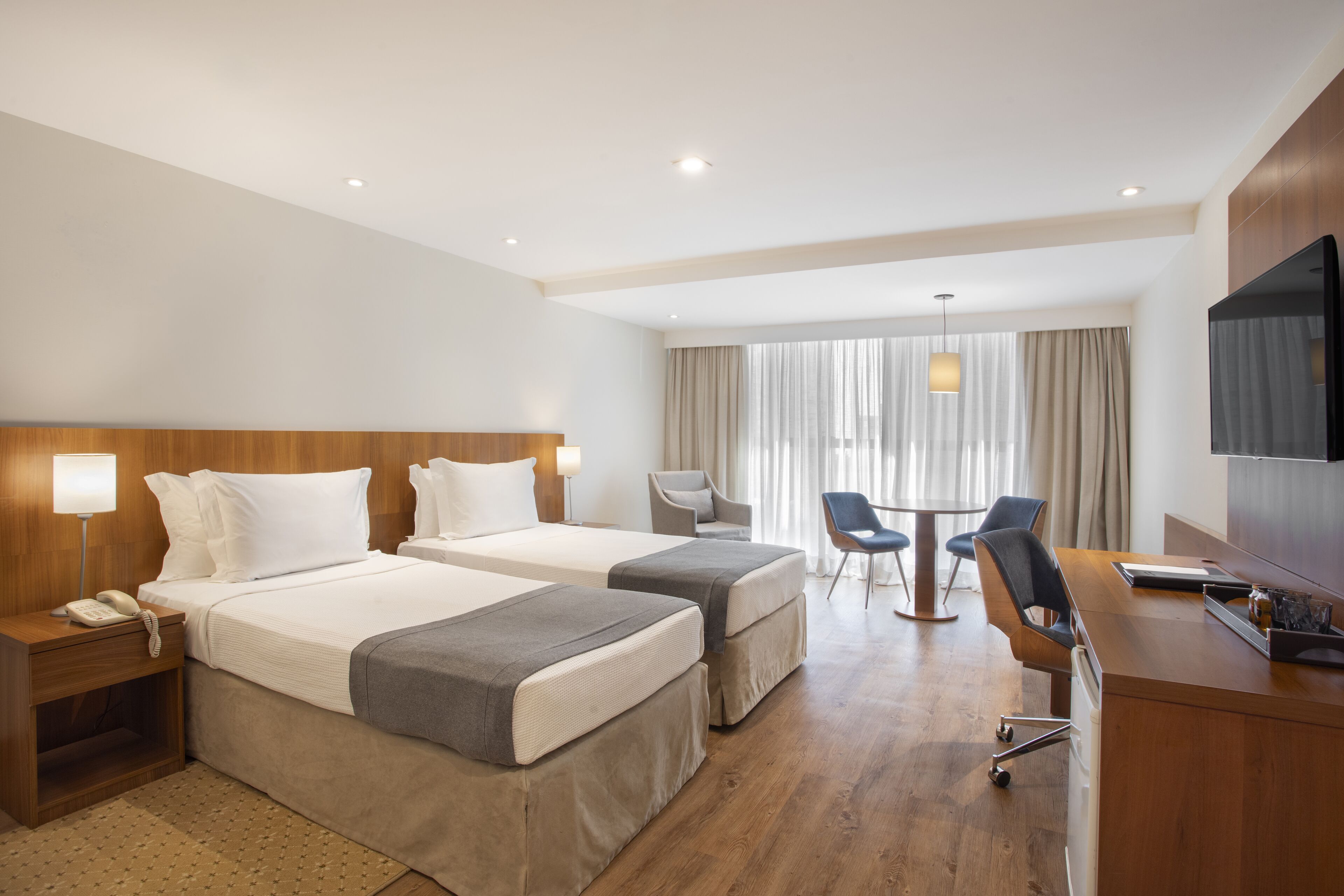 A hotel room with two beds, a desk, a chair, a TV, and a window with curtains.