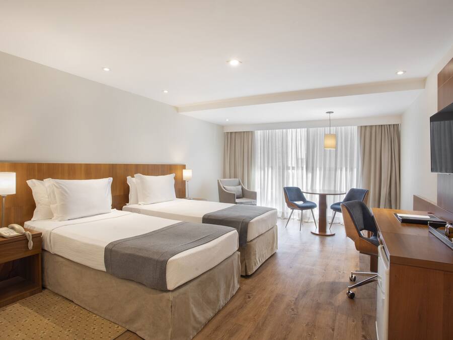 A hotel room with two beds, a desk, a chair, a TV, and a window with curtains.