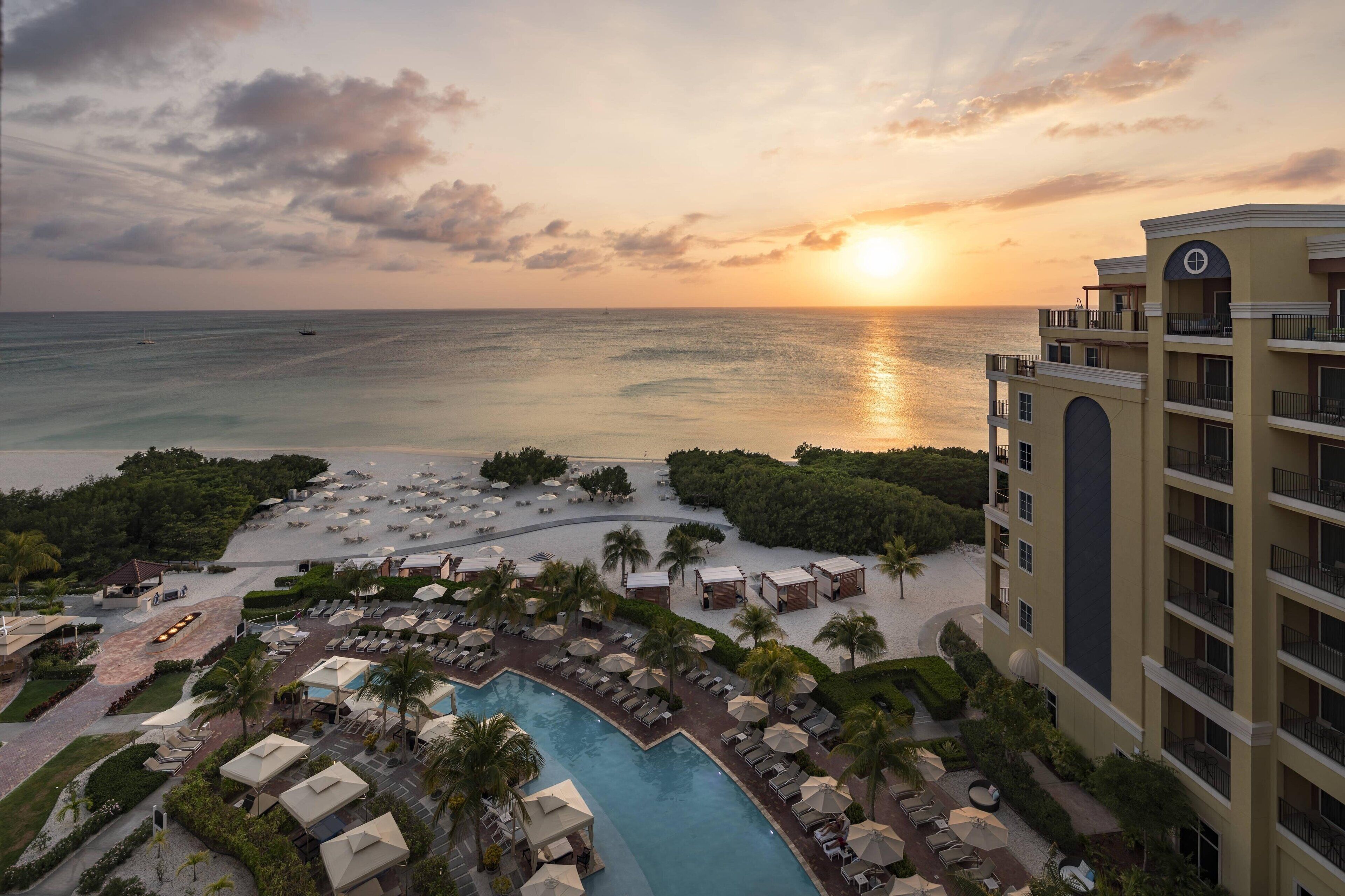 A resort with a pool, sun loungers, and a view of the ocean at sunset.