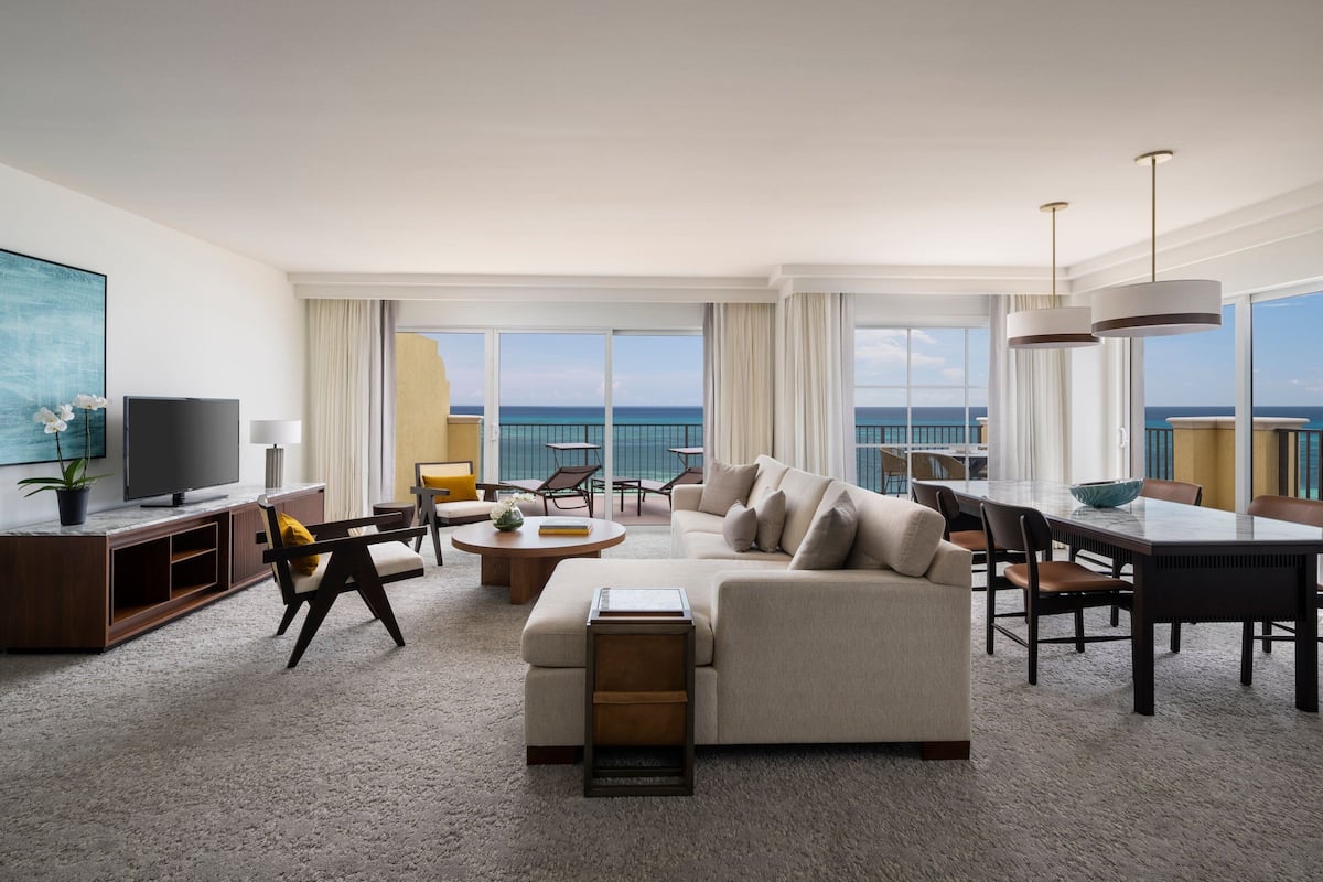 Habitación de hotel moderna con un sofá grande, una mesa de comedor y sillas. Hay un televisor sobre un mueble de madera y un ventanal con vistas al océano.