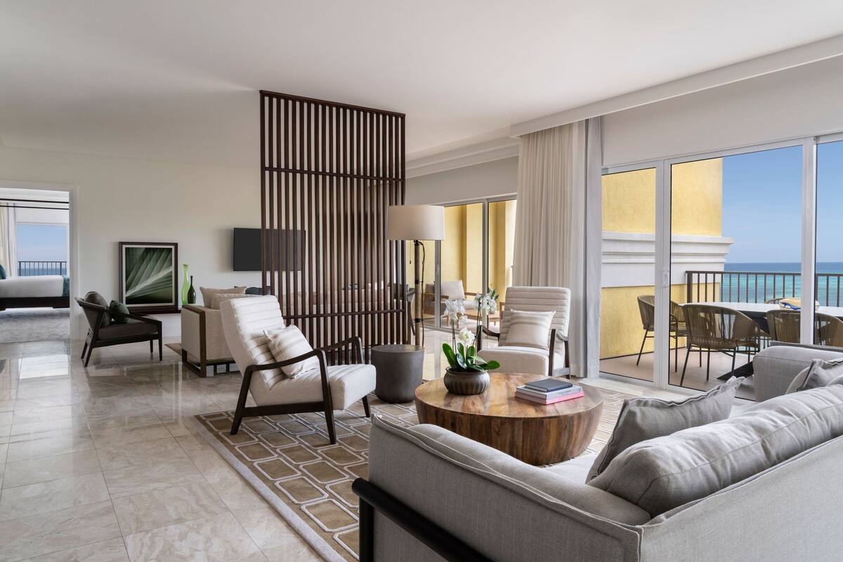 Habitación de hotel moderna con un balcón amplio, un sofá, una mesa de centro con un libro y flores, y un televisor en la pared.
