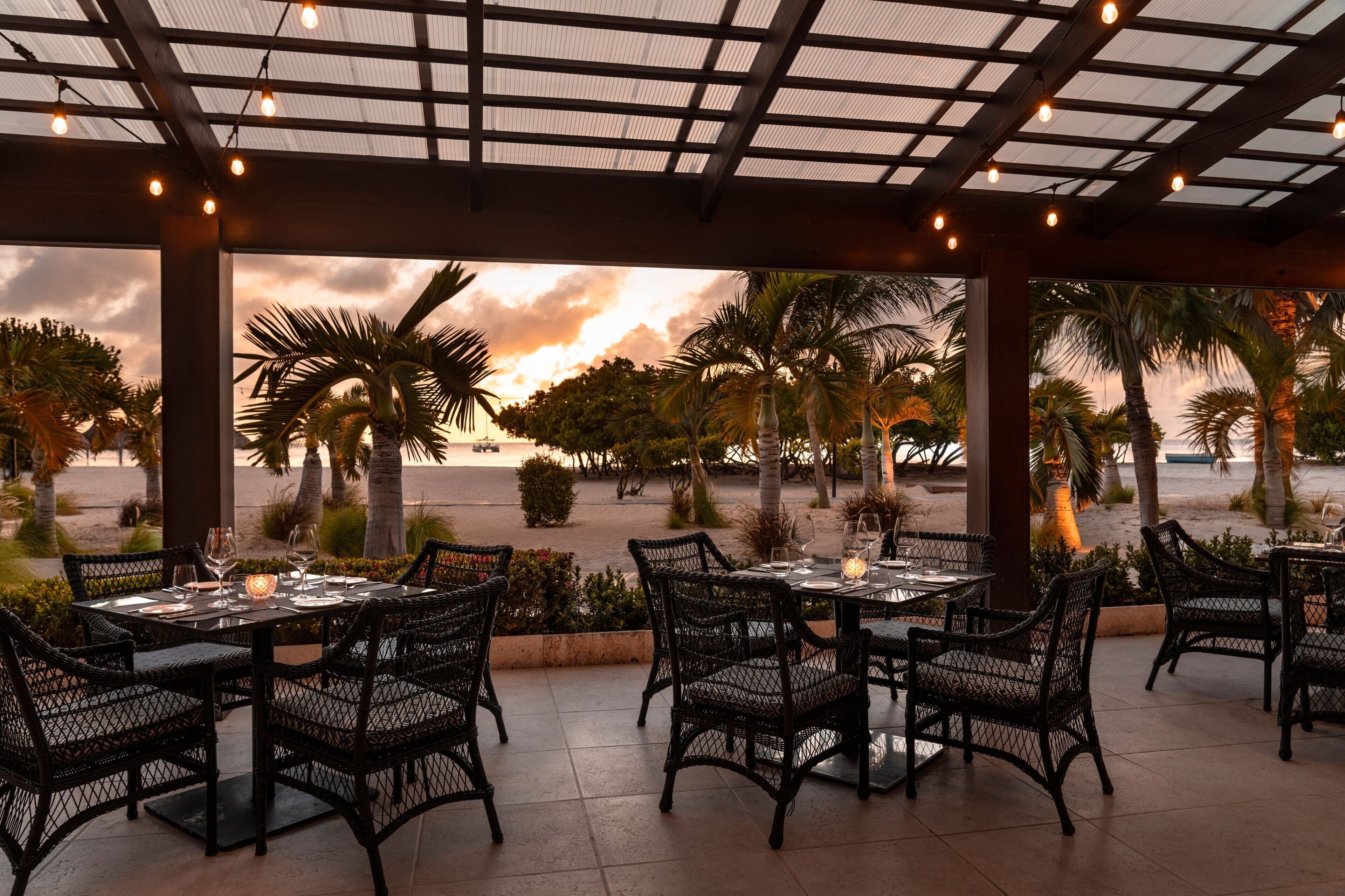 Un espacio de comedor al aire libre con mesas y sillas, con vista a una playa con palmeras y un atardecer.