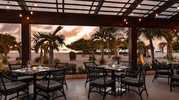 Un espace de restauration en plein air, avec des tables et des chaises, donnant sur une plage bordée de palmiers et offrant une vue sur le coucher du soleil.