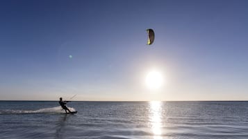 Eine Person windsurft auf dem Wasser, im Hintergrund ist ein Parasegel zu sehen.