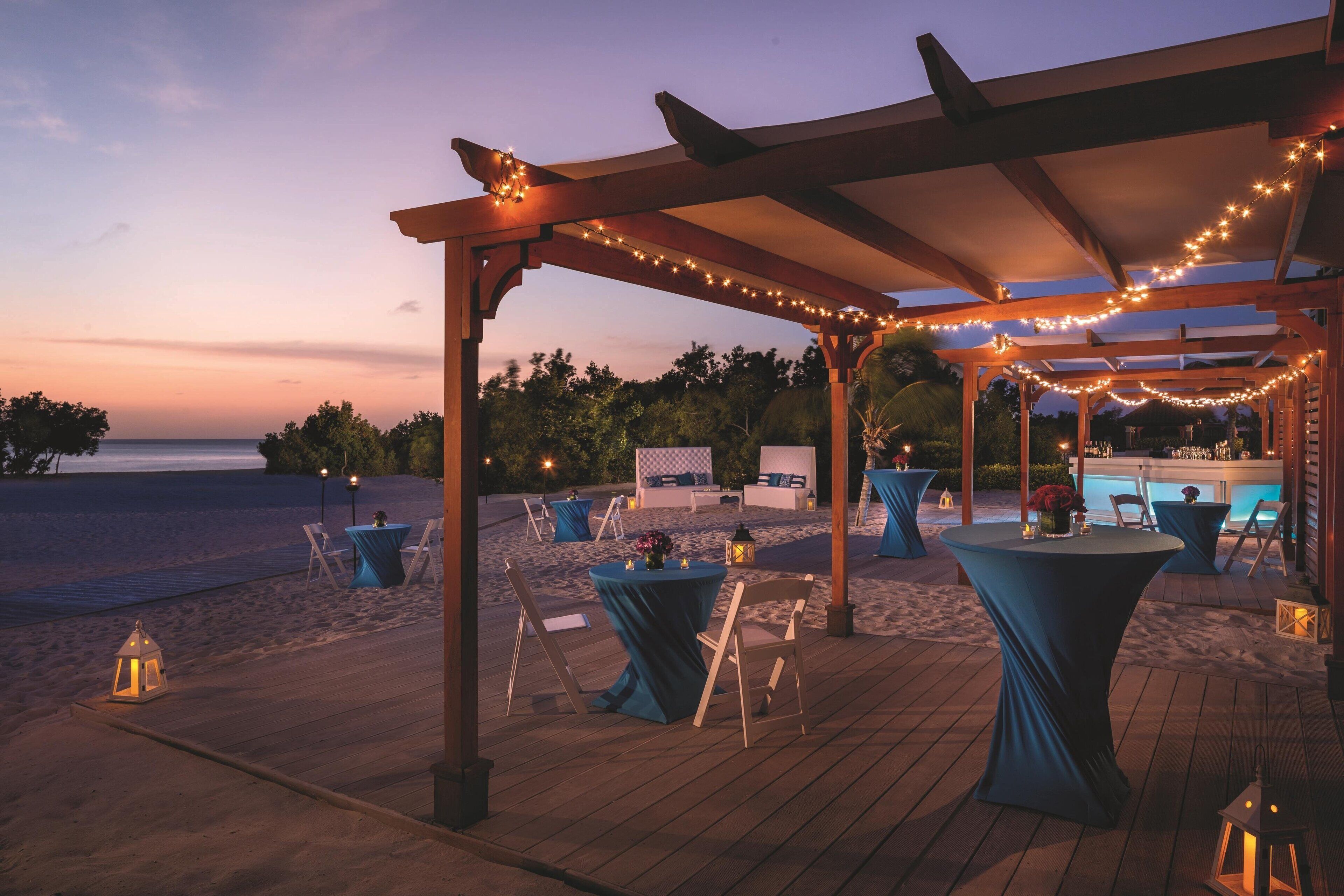 Un patio al aire libre con mesas y sillas, luces colgantes y vistas a la playa y al océano al atardecer.