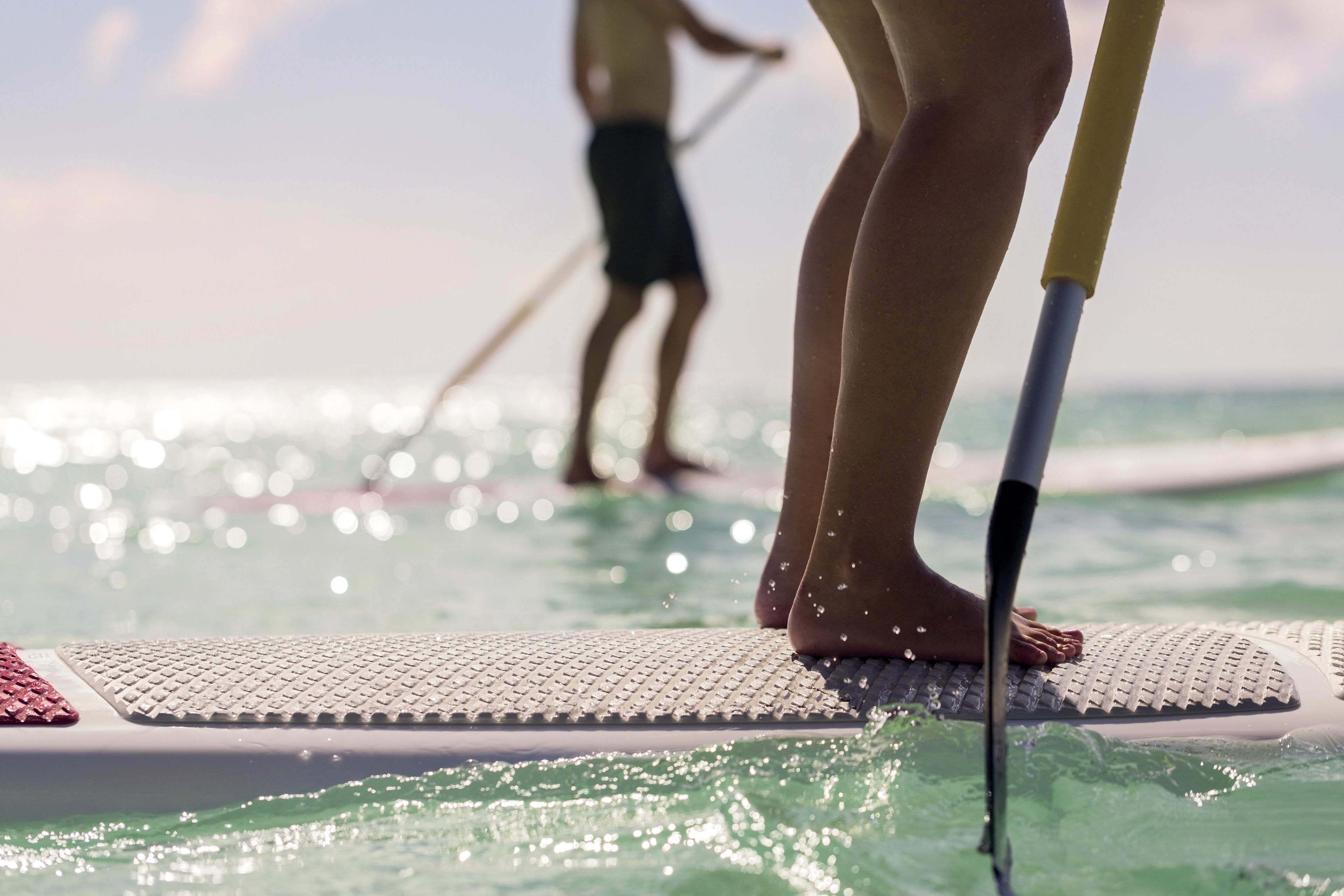 Une personne se tient debout sur une planche de paddle au milieu d’une étendue d’eau, tandis qu’une autre personne pratique le paddle en arrière-plan.