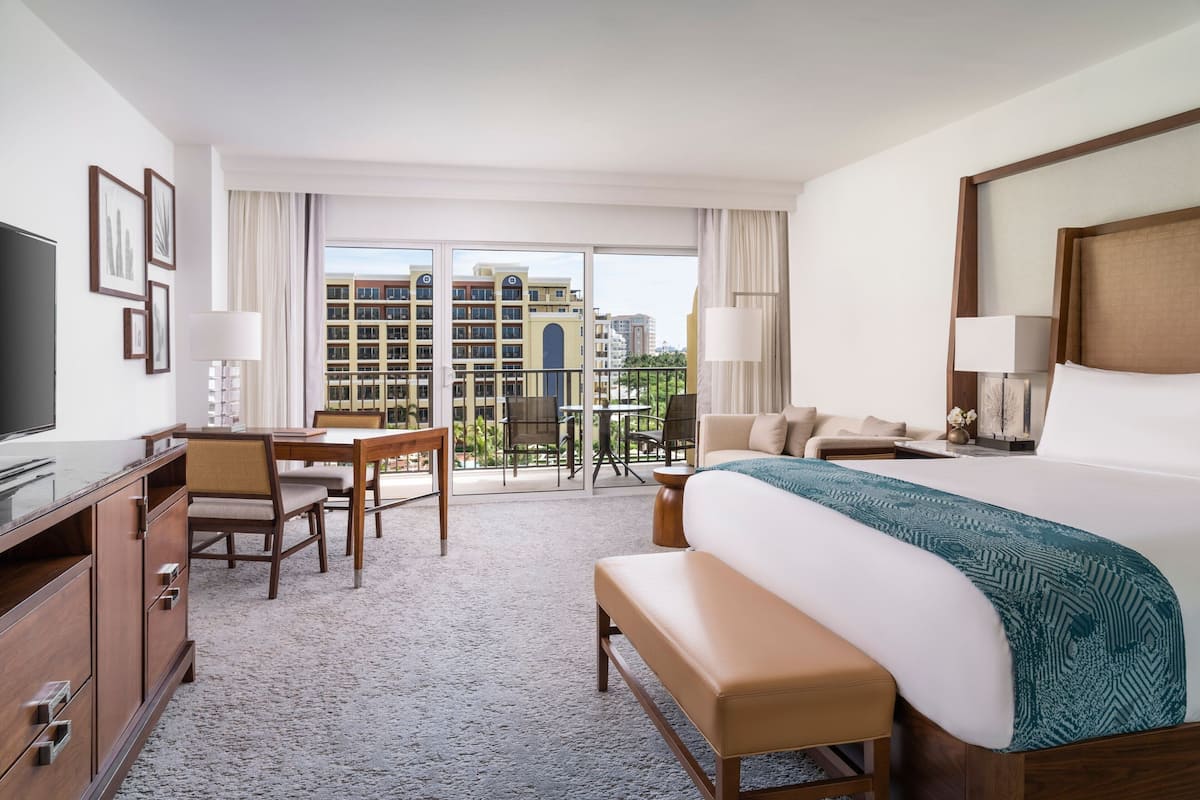 Habitación de hotel con una cama grande, un escritorio, una silla, un televisor y un balcón con vistas a la ciudad.