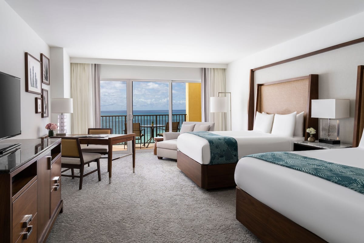 Habitación de hotel con cama, escritorio, silla, televisor y balcón con vista al océano.
