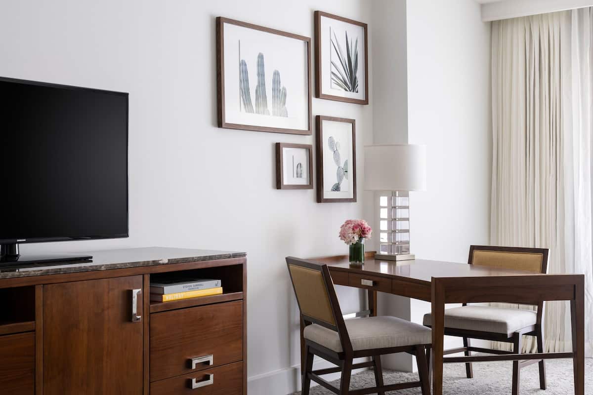 Habitación de hotel con televisor de pantalla plana, escritorio de madera, dos sillas, una mesa con un jarrón de flores, obras de arte enmarcadas en la pared y una ventana con cortinas.