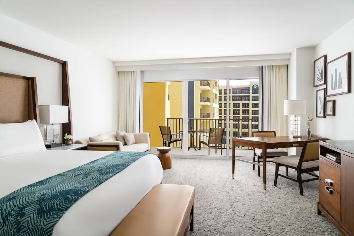 Habitación de hotel con una cama grande, un escritorio, una silla, una mesita y un balcón con vistas a edificios.