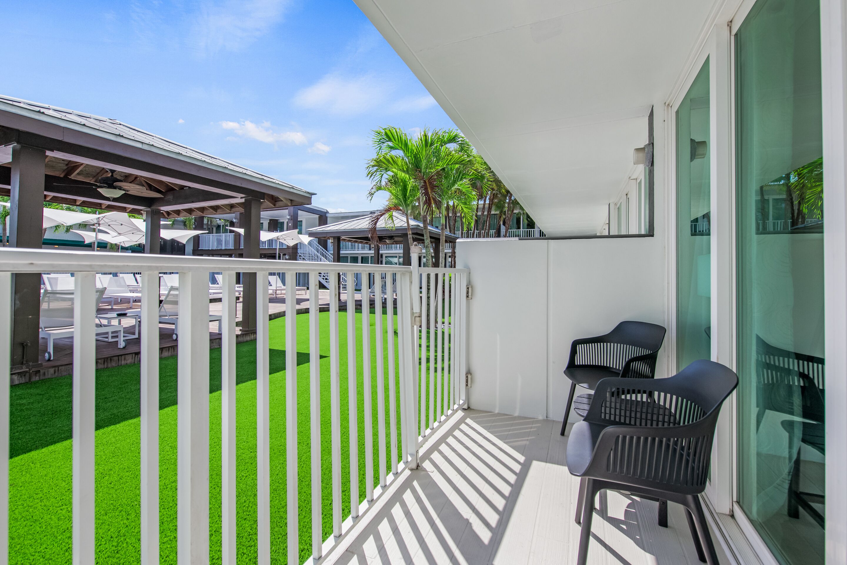 A balcony with a white railing, two black chairs, and a view of a parking area with cars and palm trees.